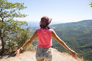 Female tourist with her hands raised looks at panoramic view on the top of the mountain and rejoices, enjoys it freedom and adventure. Trekking, travel, active ecotourism, healthy lifestyle, hiking photo