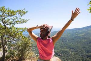 mujer con sombrero mira la vista panorámica desde la montaña hasta el mar y el bosque. turista, trekking, viajes. ecoturismo activo, estilo de vida saludable, aventura foto
