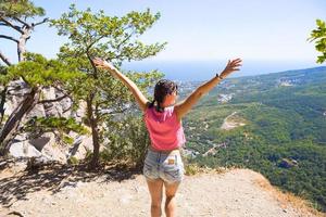 Female tourist with her hands raised looks at panoramic view on the top of the mountain and rejoices, enjoys it freedom and adventure. Trekking, travel, active ecotourism, healthy lifestyle, hiking photo