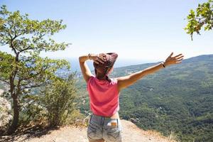 Female tourist with her hands raised looks at panoramic view on the top of the mountain and rejoices, enjoys it freedom and adventure. Trekking, travel, active ecotourism, healthy lifestyle, hiking photo