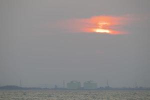 el sol rojo en el cielo y las nubes nocturnas están sobre tangyard. en la industria petroquimica .el frente es mar y olas blancas.