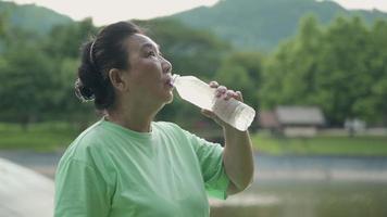 asiatische Seniorin trinkt eine Flasche Wasser nach dem Training im Park, steht draußen und entspannt sich nach dem Training, glückliches aktives Ruhestandskonzept, erfrischend nach dem Training, gesunder Lebensstil video