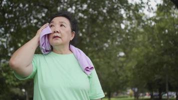 Aziatische overgewicht oudere vrouw die handdoek gebruikt om zweet af te vegen na het sporten in het park, buiten staan en rusten na het sporten, gelukkig pensioenleven, rusten na het sporten, gezondheidstoestand video