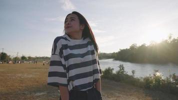 Asian girl feeling relief and relax walking along the river stream feel surrounding wind breeze, feeling the nature with arm reaching out, looking at sunset, healthy vitality breathing fresh air video