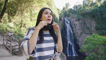 Young beautiful asian woman removing protective face mask from face, breathing oxygen in fresh air, travel destination, famous viewpoint station, holiday hiking, yelling at waterfall, emotional relief video