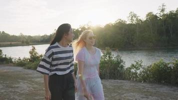 joven novia asiática lgbt pasando tiempo juntos caminando por el arroyo natural del río con el sol a punto de ponerse a la distancia, relación amorosa, ambiente cálido de primavera, dos mujeres caminan y hablan al aire libre video