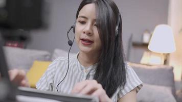 Asian young female wear headset talking on conference call, microphone voice interact, study e-learning, typing on the wireless keyboard, Online educational course,  stay at home livingroom on floor