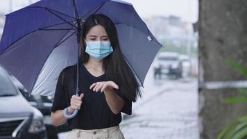 jovem mulher asiática usa máscara facial segurando guarda-chuva em pé no caminho do lado da rua, no dia chuvoso, chuveiro estação chuvosa na ásia, carro estacionado na estrada, estenda o braço pegando gotas de chuva