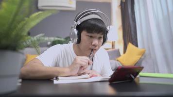 Asian college boy watching lesson online and studying from home. Young man taking homework notes while looking at smartphone screen, isolated e-learning, coz comfortable living room, human knowledge video