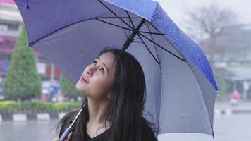 A young beautiful asian woman stands on roadside with holding a blue umbrella on rainy day caused by a tropical typhoon, waiting for bus on city roadside, weather forecasting, cold and flu prevention video