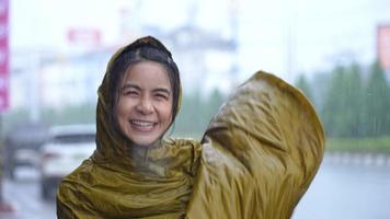 Young cheerful woman in yellow wet raincoat jump and dancing on footpath next to main road with cars moving, having fun among heavy rain on raining day, funny girl dancing, living in rainy season