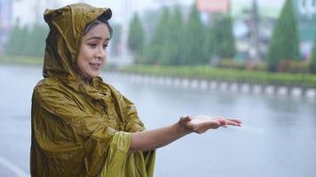 Young tired female office worker in yellow raincoat stuck in a rain, stands under roof and playing with hands collecting raindrops, heavy rain caused by monsoon, asian city life, tropical zone country video