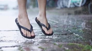 Low angle shot feet in flip flops stomping on water puddle on rainy day caused by monsoon, enjoys jumping and splashing water droplets around paving walkway, slow motion, tropical climate changes video