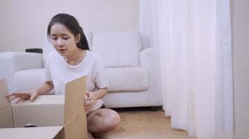 Young adult asian woman unpack  carton parcel box, looking at old stuff arranging sorting things, sit down in the living room, good old memories, Relocation moving into new apartment, front view video