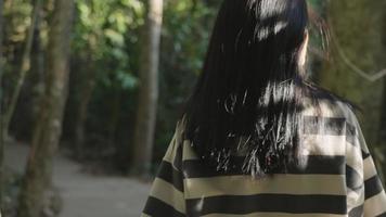 vue arrière photo d'une jeune femme asiatique active marchant dans la forêt tropicale avec la lumière du soleil qui brille progressivement à travers son visage, une activité de sentier de randonnée d'aventure, de longs cheveux noirs en soie qui brillent, un environnement vert video