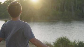 Young asian man feeling relief and relax standing looking at water river flows, arms reaching out feel surrounding wind breeze, exploring feeling the nature destination, sun flares warm summer weather video