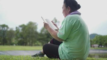 femme âgée asiatique s'asseoir pour se reposer et lire un livre dans un parc extérieur après l'exercice, activité de loisirs de la vieillesse, vie de retraite, relaxation de la vitalité du bien-être, lectrice obèse sur une herbe confortable video