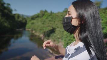 câmera lenta de uma jovem e linda garota asiática com remover a máscara facial e respirar profundamente em pé na ponte pelo córrego do rio apreciar a beleza e a pureza da natureza, área aberta ao ar livre durante a pandemia