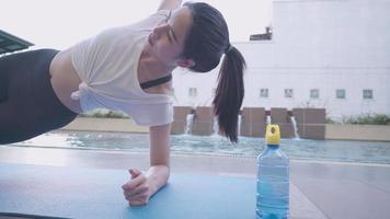 actieve jonge aziatische vrouw doet zijplank op oefenmat voor sterkere buikspieren zes packs kern, zelfmotivatie discipline, fitnesstraining in sportkleding, condominium openluchtfitnessclub zwembad