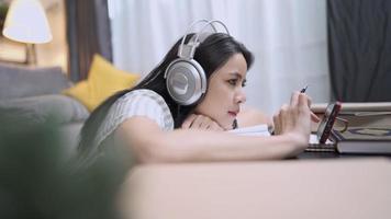 Aziatisch meisje met een headset die zich lui voelt rusten door online entertainment op het smartphonescherm thuis in de woonkamer te kijken, online les, zelfstudie op afstand, naar muziek luisteren video