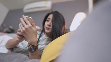 joven mujer asiática que usa un teléfono inteligente acostado en un cómodo sofá, compras en línea, conexión inalámbrica wifi en casa, relajación sin preocupaciones en el tiempo libre el fin de semana, sala de estar simple vida inteligente