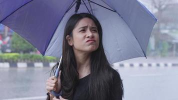 jeune femme asiatique aux longs cheveux noirs déçue coincée sous une pluie battante à l'extérieur, main utilisant un parapluie pour éviter de se mouiller, expression faciale de sentiment négatif, changements climatiques tropicaux video