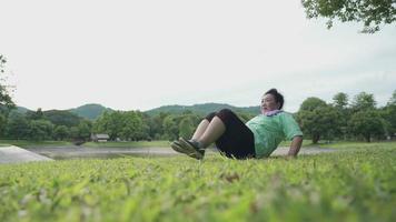 donna anziana obesa attiva asiatica che fa esercizi addominali a terra, alberi di campo di erba anziani, femmina di mezza età che si esercita al parco all'aperto, vita di vecchiaia che diventa forte, telecamera grandangolare video