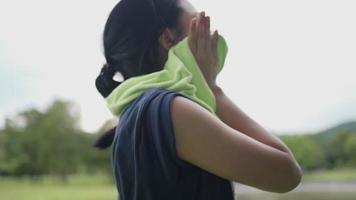 Young asian woman using towel on neck wiping sweat on face and neck during exercise at green park, take a break from training, woman drying sweat, tiredness and sport concept, active healthy lifestyle