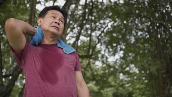 homme asiatique d'âge moyen essuyant la sueur sur son visage et son cou pendant l'exercice au parc, debout sous les arbres et se reposer après l'exercice, bien-être du corps fatigué épuisé, état de santé médical, angle bas video