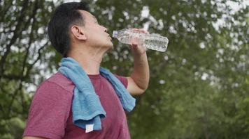 asiatischer älterer mann trinkt wasser während der pause während des trainings im park mit bäumen im hintergrund, steht im freien und erfrischt sich nach dem training, glücklicher aktiver pensionierter lebensstil video