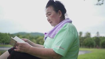 Aziatische senior vrouw gaat zitten en leest een boek in het buitenpark na het sporten, vrijetijdsbesteding op oudere leeftijd, pensioenleven, wellness-vitaliteitsontspanning, zwaarlijvige vrouwelijke lezer op comfortabel gras video