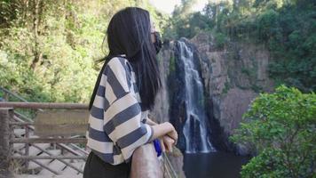 A young asian female tourist wears medical mask, stands on viewpoint station in front of national park's waterfall, travelling in the new normal, social distancing, prevent spreading Covid-19 Delta video