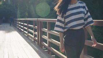 A female body in casual wear walking on a wooden bridge inside natural park, crossing a river, a long straight black hair flowing by fresh wind, touching natural rainforest vibe, hot summer vacation video