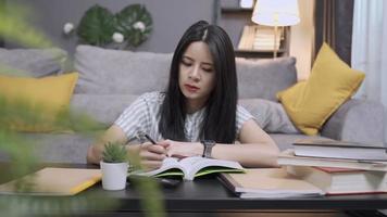 une fille asiatique s'assoit pour étudier seule dans le salon confortable de la maison, écrivez des idées sur les devoirs papier, asseyez-vous sur le bureau à domicile au sol, auto-apprentissage de l'éducation à domicile pendant la quarantaine de verrouillage video