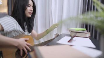 Young attractive asian girl learning guitar lesson for beginner according to online teaching app, learning or improving hard skills, leisure activity at home during COVID-19, relaxing sing and song video