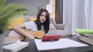 mooi Aziatisch meisje dat akoestische gitaar speelt in de woonkamer van het huis, harde vaardigheden leren of verbeteren, vrijetijdsbesteding thuis tijdens covid-19, ontspannend zingen, leren hoe je een modern instrument bespeelt video