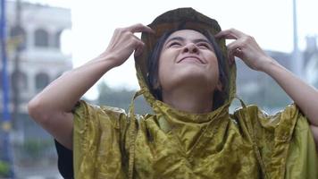 una chica asiática se quita la capucha impermeable para disfrutar de la lluvia que cae, parada al lado de la carretera, mirando hacia el cielo disfrutando de las gotas de lluvia en su cara sonriendo, sintiéndose fresca, cambiando el clima de la temporada video