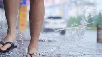 A flip flops feet carefully running on an urban paving footpath during a day, low angle shot on female legs in casual clothing style, a car parked on street road, splashing purity water puddle