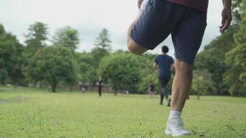 asiatiska manliga stretch benmuskler innan jogging träning inne i parken med träd och grönt gräs och människor i bakgrunden, kroppskonditionering värm upp kroppen innan du springer jogging, flexibelt ben video