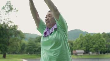 Asian mature woman doing over head arm stretching before exercise in the outdoor park with trees mountain background, morning exercise routine, active elderly aged woman, retirement life stronger body