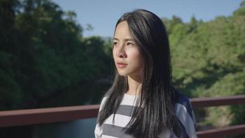 attraente turista dai capelli neri che cammina lentamente da solo sul ponte di legno mentre attraversa il fiume mentre fa un'escursione all'interno di una foresta pluviale tropicale, con la luce solare che splende sul viso femminile contro il fogliame lussureggiante video