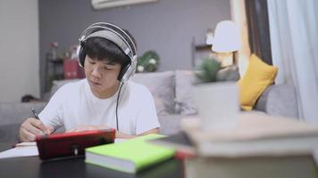 A young asian man doing self study at home living room on weekend, casual cozy style, distant online learning by using smartphone, active student put on headphone and writing note on paper book video