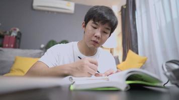 Asian male student studying inside house living room, sit down on floor with couch behind, doing homework, home education Online learning, distance lesson, education at home, focusing on study video