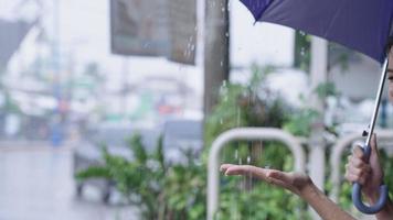 una joven y hermosa mujer asiática alcanzando el brazo para recoger algunas gotas de agua del paraguas que sostiene, una trabajadora infeliz y preocupada que llega tarde al trabajo, desesperada sentada en el sendero de la calle, temporada de lluvias video