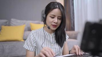 mujer joven asiática usa auriculares con micrófono interactúa hablando llamando con la base de datos de búsqueda de voz, estudia e-learning, escribe en el teclado inalámbrico, trabajador independiente en línea, acogedora sala de estar video