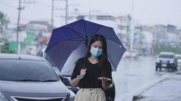 ung asiatisk kvinna med svart hår går utanför bland ett kraftigt regn orsakat av tropisk tyfon, avslappnad flicka känner sig trött på att bli blöt, håller paraplypromenad på stadsgatans gångstig med bilparkering bakom video