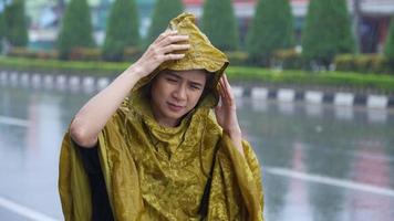 An east asian woman wearing raincoat raising hand checking on level of pouring rain while walking on street footpath, rainy season preparation, tropical climate change, getting wet in rain, video