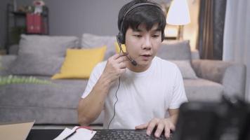 young adult male Asian wear headset microphone doing calling operator taking notes from client information, microphone voice call interaction with customer, live chat with staff doing working at home video