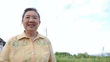 uma mulher madura do leste asiático confiante andando olhando para a câmera, sorriso gesticulando, exercício relaxante na rua da vila, idosas passeando em um dia de bom tempo com um céu branco claro