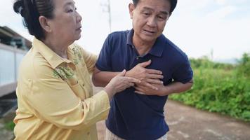 Senior asian man suddenly feeling the pain in his chest, having heart attack while walking outside house neighborhood with his wife along side, health insurance, pain in the chest, medical emergency video